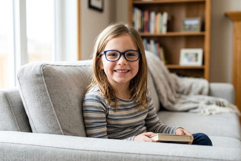 Child in blue glasses and striped shirt smiles while sitting on a grey sofa holding a book, bookshelf in the background.